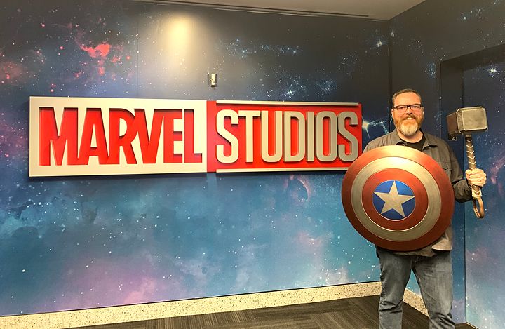 Mike McHargue grinning in front of the Marvel Studios sign, holding Captain America's shield in one hand and Thor's hammer in the other.