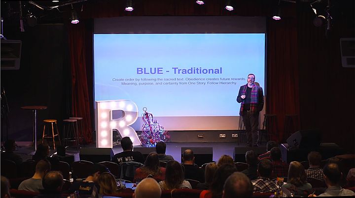 Mike McHargue on stage in a Dublin theater, speaking beside a large projected slide while a seated audience watches.