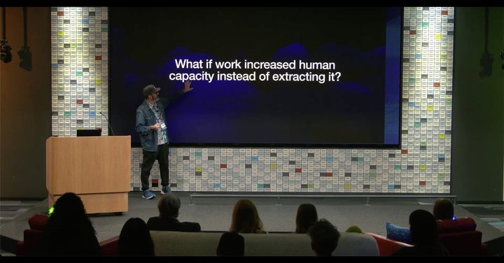 Mike McHargue speaking at a podium during Google Xi Days 2026, addressing a seated audience in an auditorium. A large projected slide behind him reads "What if work increased human capacity instead of extracting it?"